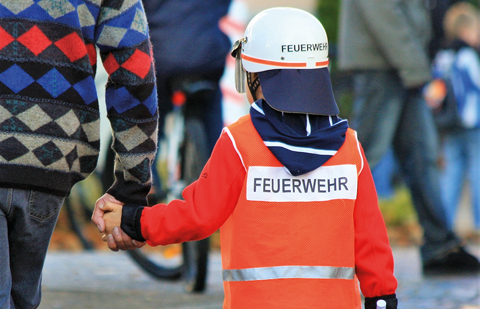 Kind mit Feuerwehrhelm und -jacke an der Hand eines Erwachsenen