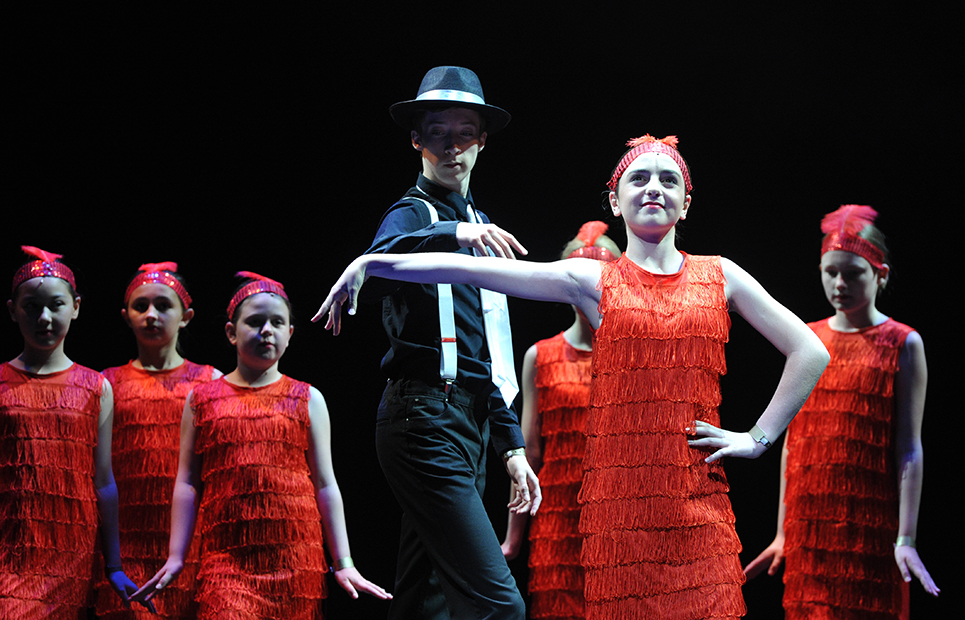 1 Junge im Anzug mit Hut und Mädchen in roten Fransenkleidern bei einer Tanz-Aufführung auf einer Bühne