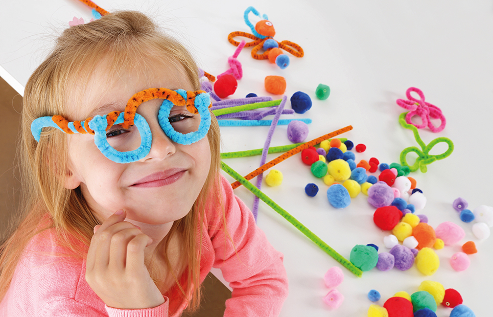 Ein Mädchen mit einer Brille aus Pfeifenputzern auf der Nase. Im HIntergrund weitere Pfeifernputzer und Pompons zum Basteln