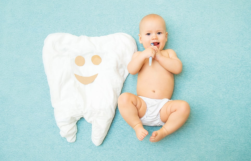 Ein Baby liegt auf dem Rücken mit einer Zahnbürste im Mund. Neben ihm liegt ein übergroßes Kissen in Zahnform.